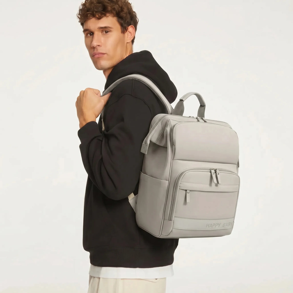 Jeune homme portant un sac à dos à langer gris clair avec double zip argenté 
et logo Happy Baby, vue 3/4 en studio fond blanc