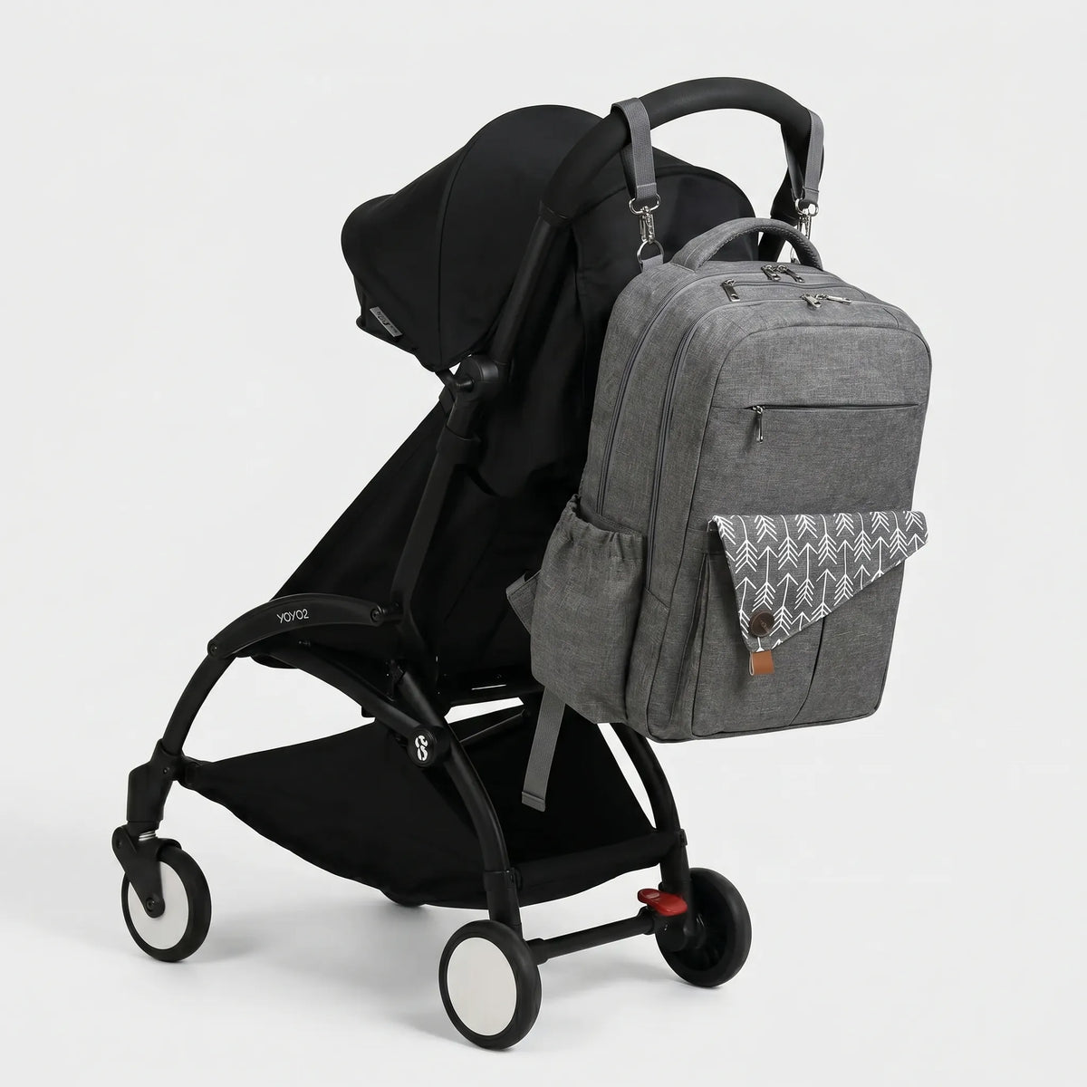 Sac à dos à langer gris anthracite avec rabat imprimé flèches blanches et sangles 
de fixation poussette, suspendu au guidon d'une poussette Babyzen YOYO2 noire — 
vue lifestyle studio