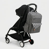 Sac à dos à langer gris anthracite avec rabat imprimé flèches blanches et sangles 
de fixation poussette, suspendu au guidon d'une poussette Babyzen YOYO2 noire — 
vue lifestyle studio