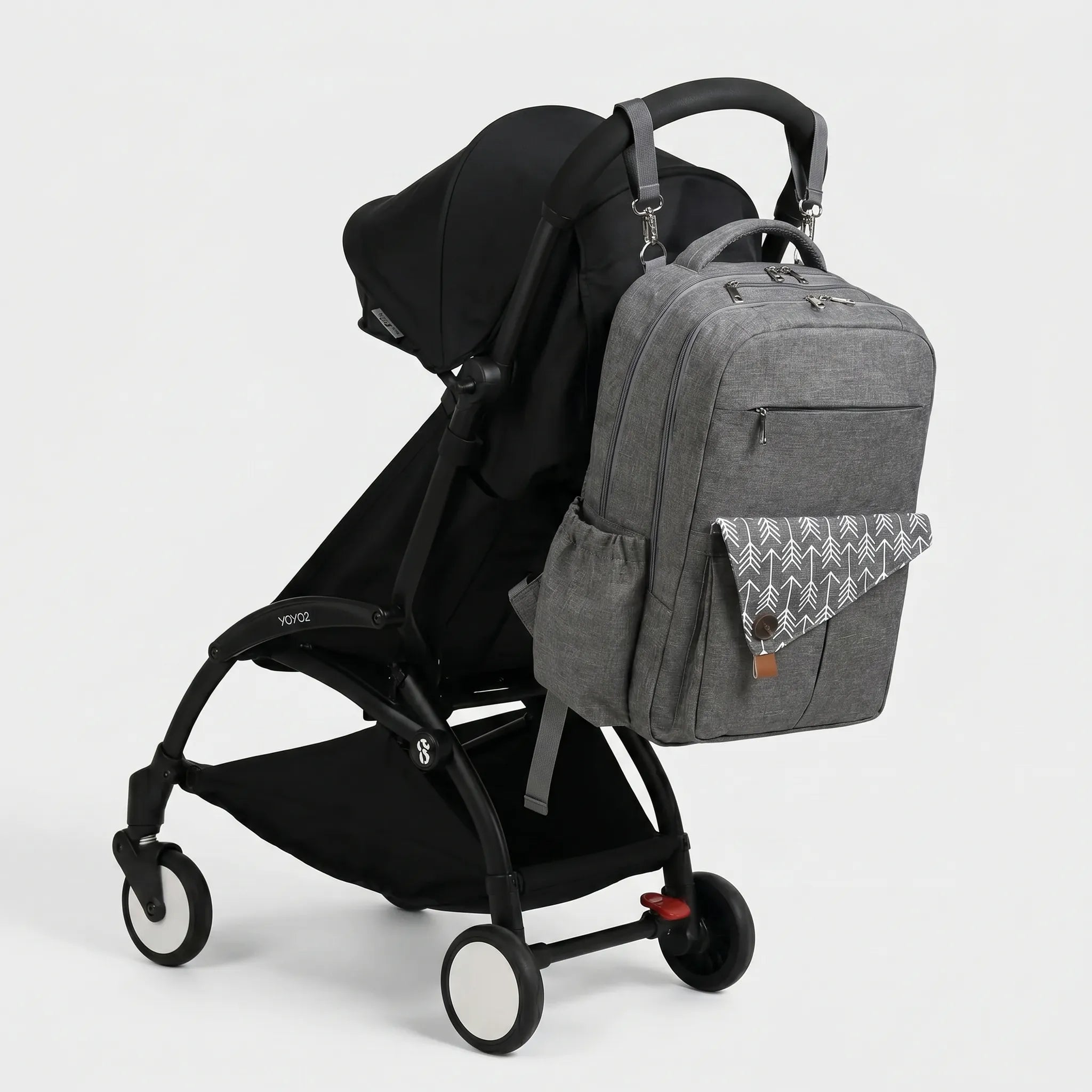 Sac à dos à langer gris anthracite avec rabat imprimé flèches blanches et sangles 
de fixation poussette, suspendu au guidon d'une poussette Babyzen YOYO2 noire — 
vue lifestyle studio