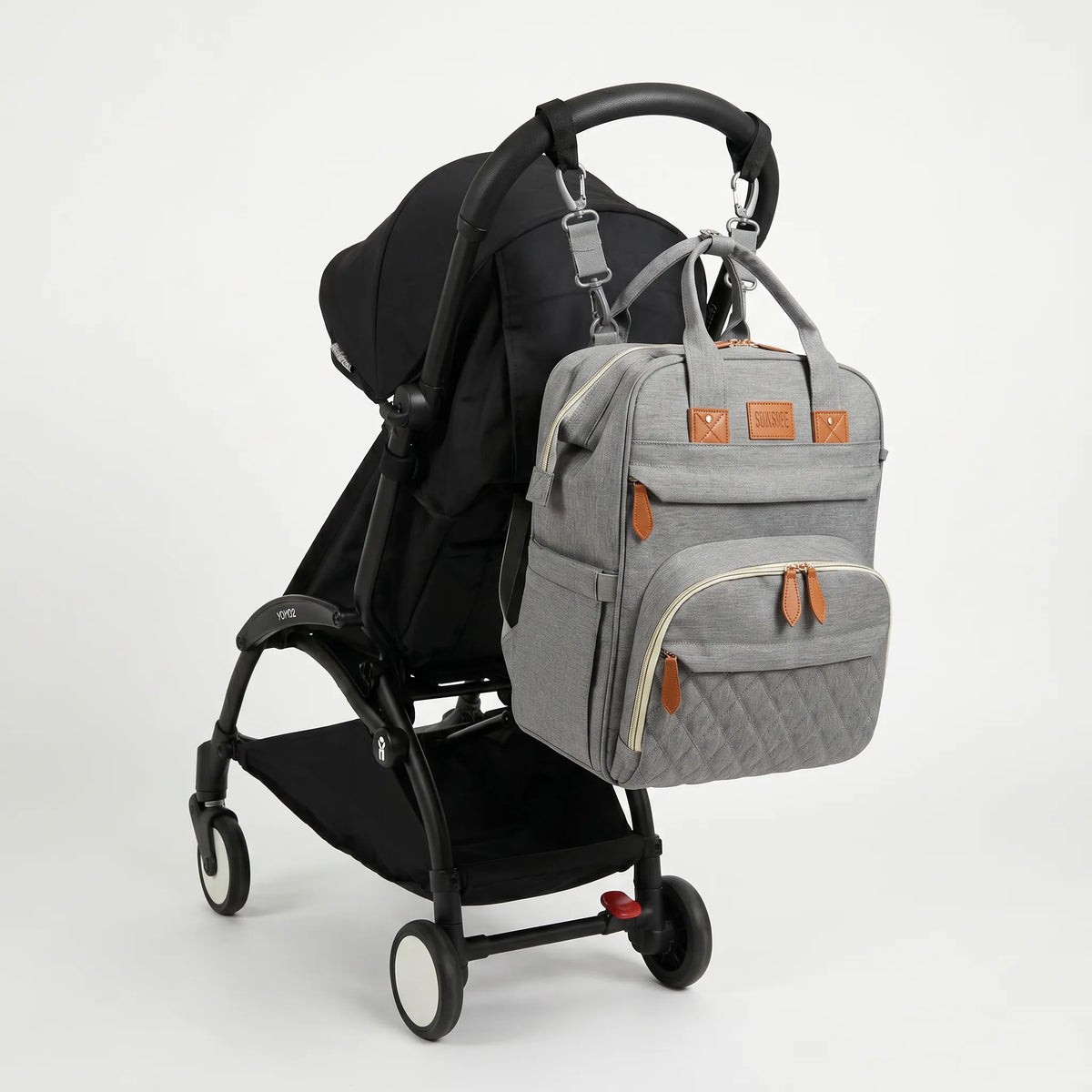 Sac à dos à langer gris chiné avec port USB intégré et accents cuir caramel, 
suspendu au guidon d'une poussette Babyzen YOYO2 noire — vue lifestyle studio