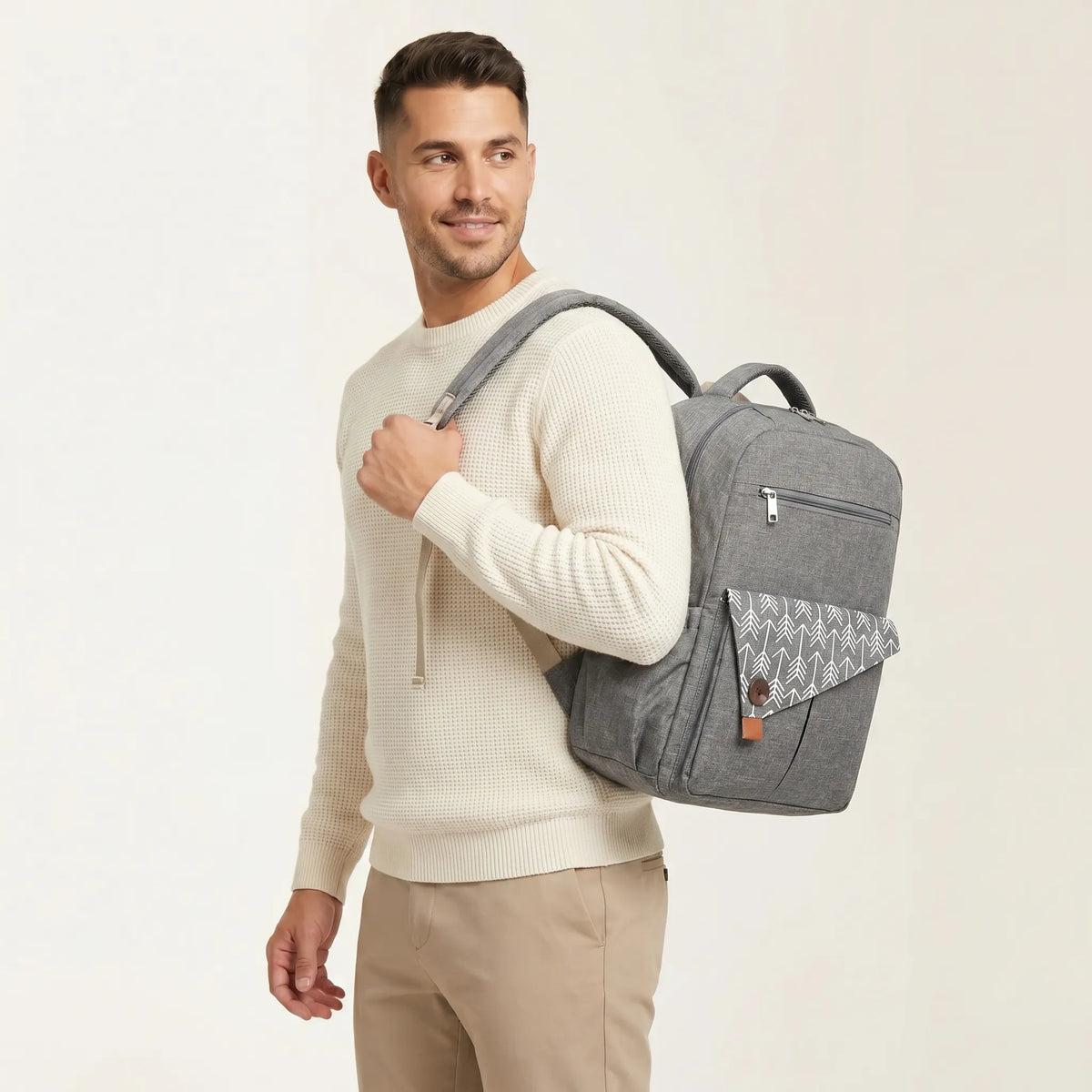 Homme aux cheveux courts en pull waffle crème portant un sac à dos à langer gris chiné avec rabat à motif flèches blanches et bouton bois sur fond studio