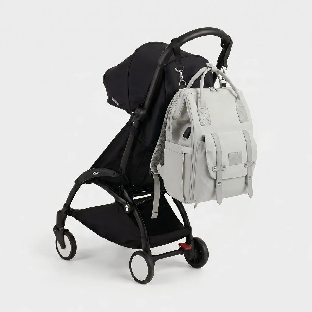 Sac à dos à langer gris clair style utilitaire avec port USB intégré et sangles 
à boucles argentées, suspendu au guidon d'une poussette Babyzen YOYO noire — 
vue lifestyle studio