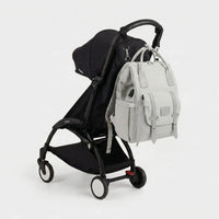 Sac à dos à langer gris clair style utilitaire avec port USB intégré et sangles 
à boucles argentées, suspendu au guidon d'une poussette Babyzen YOYO noire — 
vue lifestyle studio