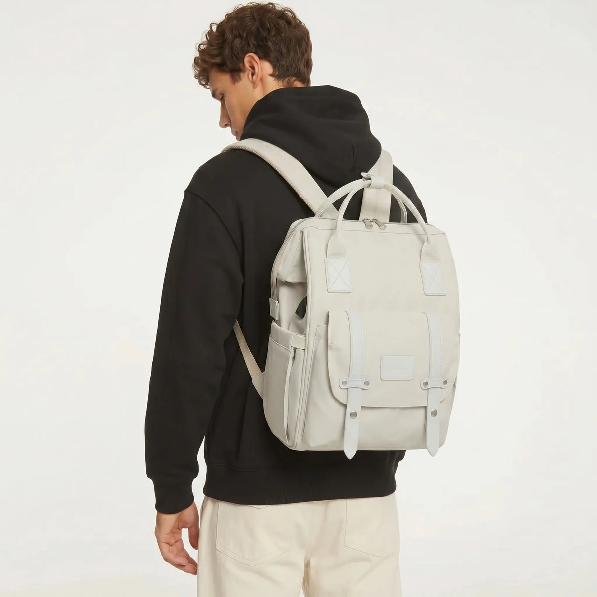 Jeune homme portant un sac à dos à langer gris clair style utilitaire avec sangles 
militaires et hardware argenté, vu de dos en studio fond blanc