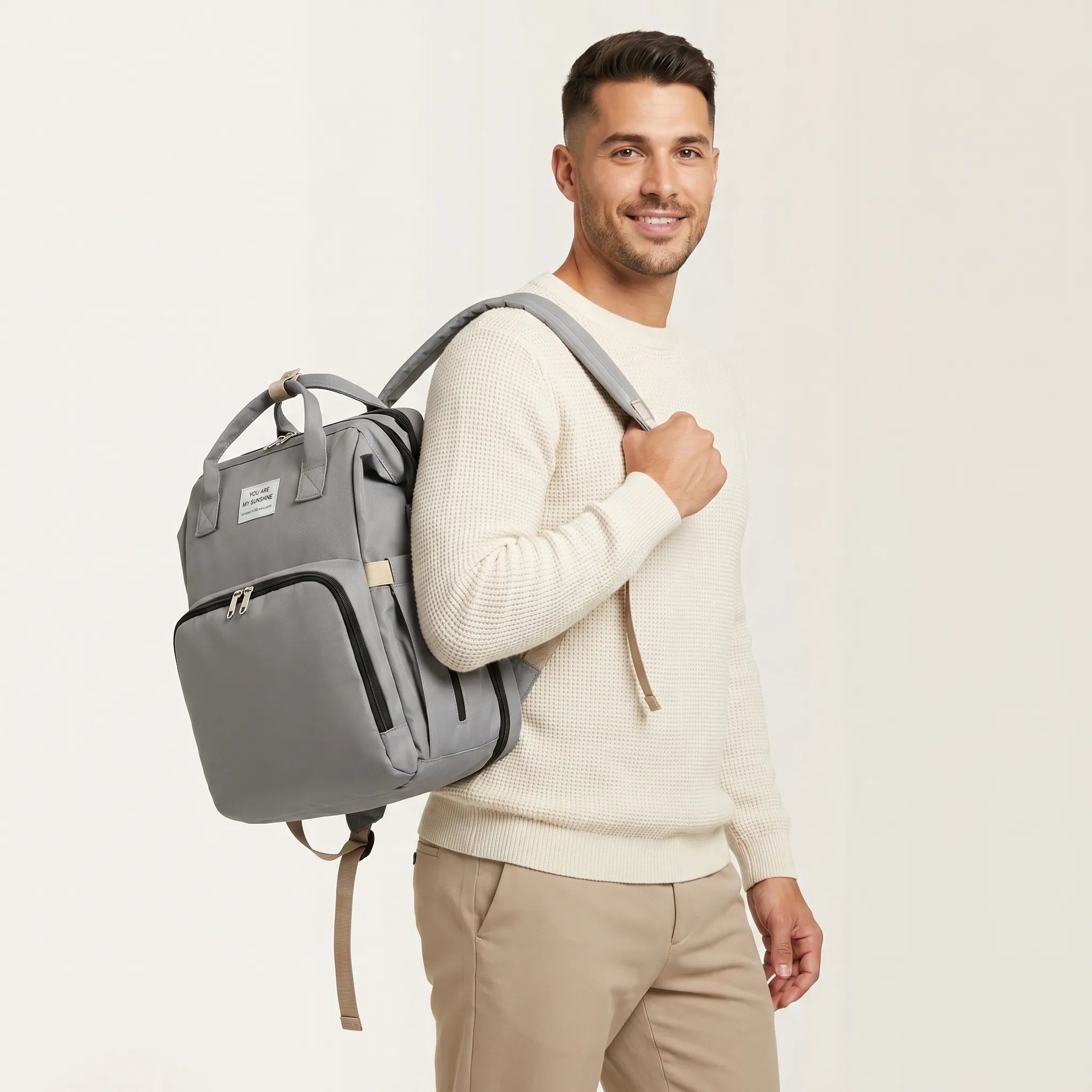 Homme souriant en pull crème portant un sac à dos à langer gris sur une épaule avec étiquette You Are My Sunshine