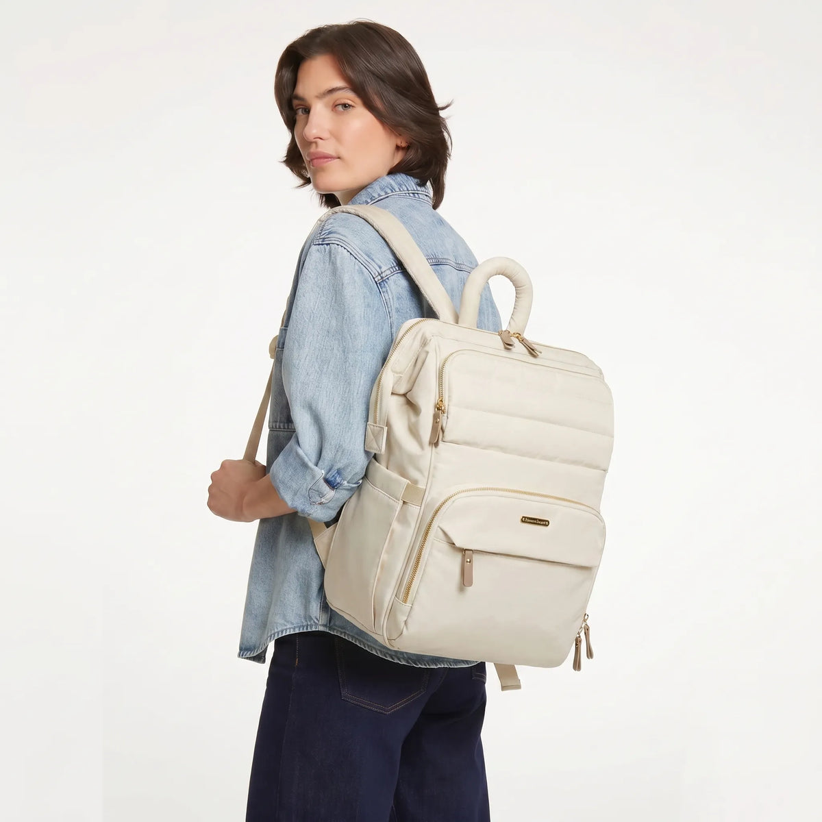Femme aux cheveux bruns en bob portant une chemise en jean clair et un jean indigo, vue de trois quarts dos avec un sac à dos à langer matelassé crème ivoire à hardware doré sur l'épaule