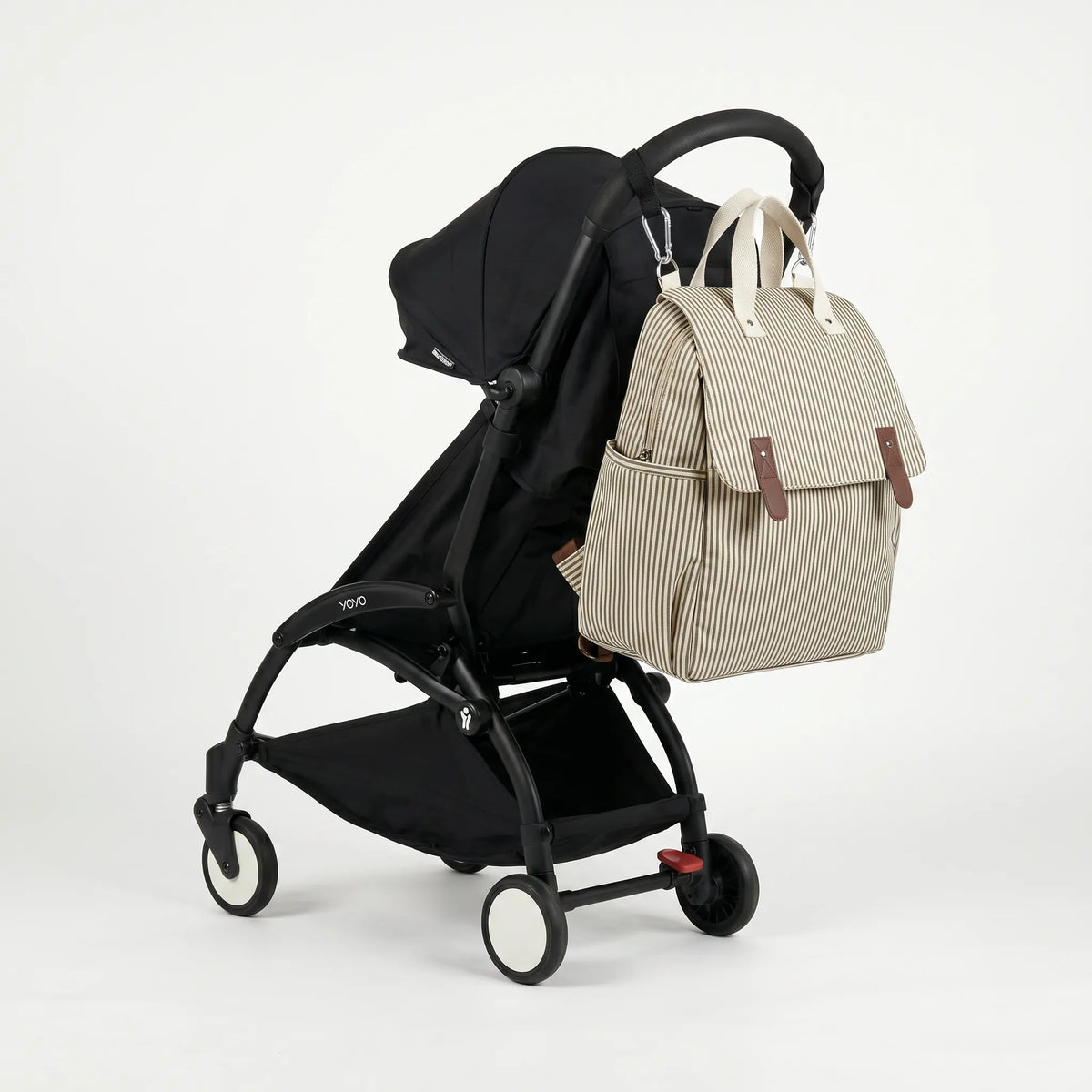 Sac à dos à langer rayé crème et kaki avec rabat et fixation poussette intégrée, 
suspendu au guidon d'une poussette Babyzen YOYO noire — vue lifestyle studio