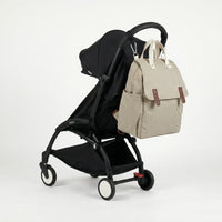 Sac à dos à langer rayé crème et kaki avec rabat et fixation poussette intégrée, 
suspendu au guidon d'une poussette Babyzen YOYO noire — vue lifestyle studio