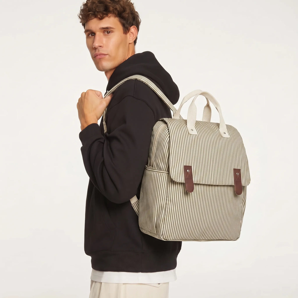 Mannequin homme aux cheveux courts bouclés en hoodie noir portant en mode single-strap un sac à dos à langer rayé écru et vert olive avec rabat à lanières cuir marron
