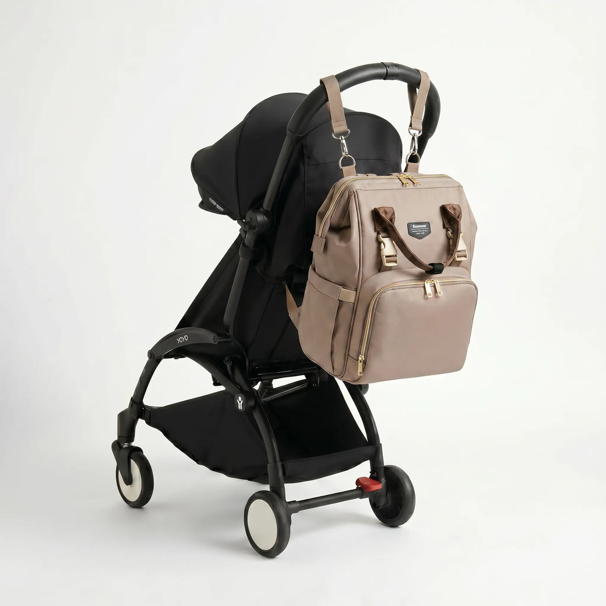 Sac à dos à langer taupe avec zips dorés, anses marron et clips de fixation poussette accroché au guidon d'une poussette compacte noire de type Yoyo