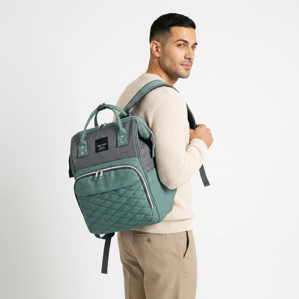 Homme aux cheveux courts portant un sac à dos à langer vert sauge et gris chiné avec motif matelassé sur fond blanc studio