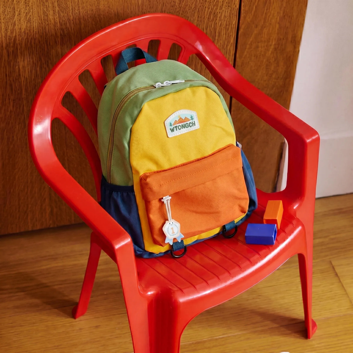 Sac à dos maternelle 2-6 ans au design colorblock vert moutarde orange et bleu posé sur une chaise enfant rouge avec cubes de motricité