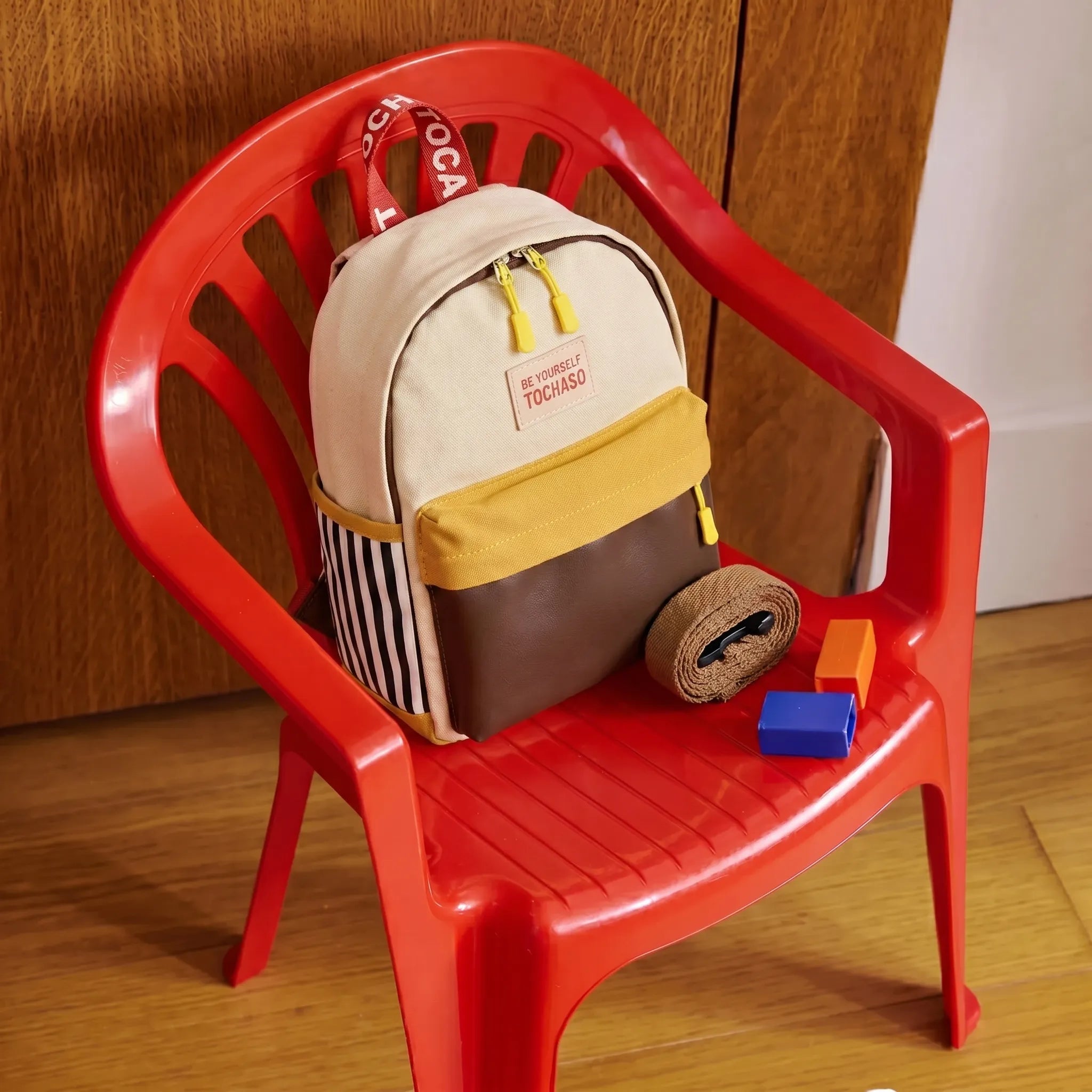 Sac à dos maternelle en toile canvas écru avec poche moutarde et cuir marron posé sur une chaise enfant rouge avec sangle de sécurité enroulée et cubes de motricité