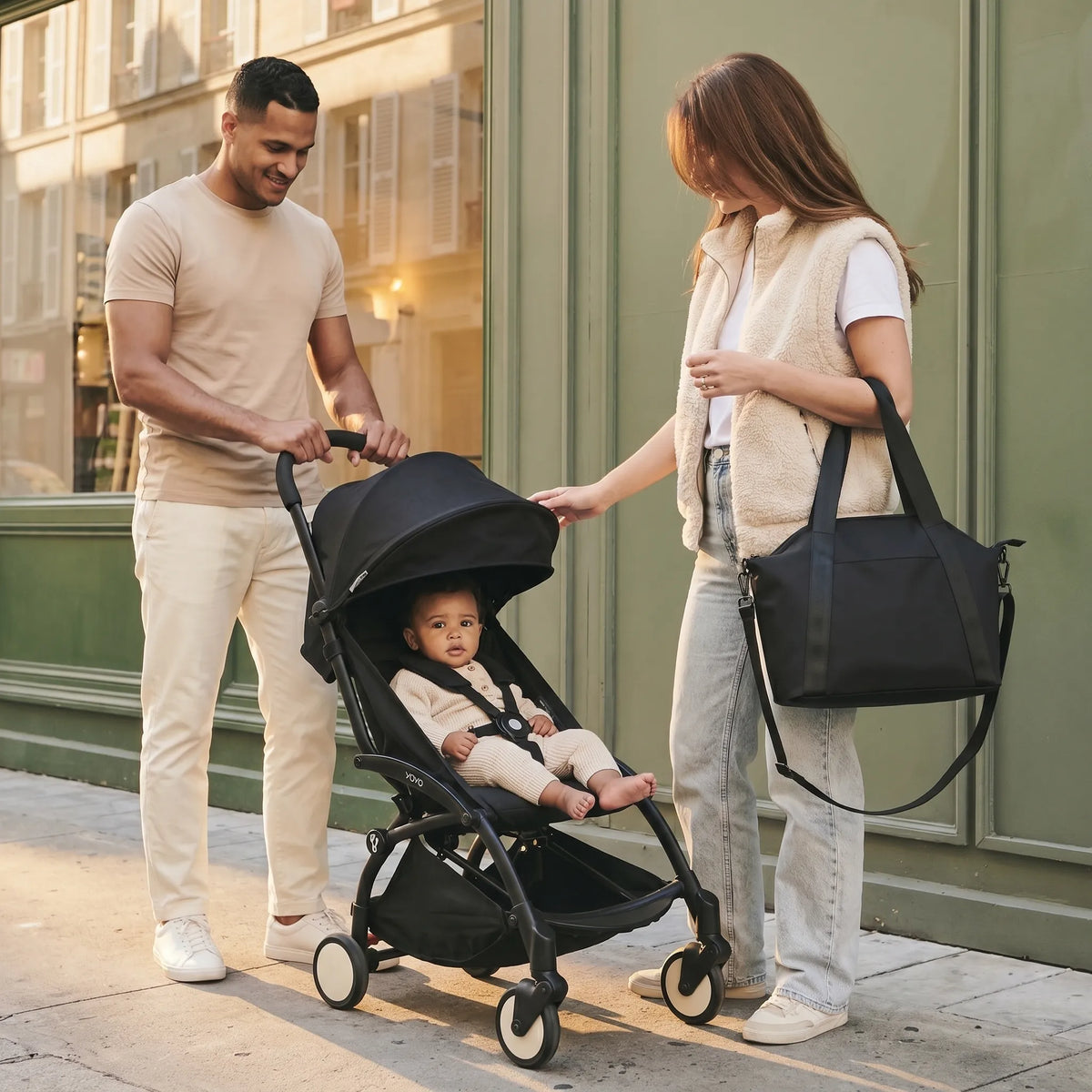 Famille parisienne moderne, papa souriant qui pousse une poussette YOYO dépliée avec bébé métis assis dedans et maman aux longs cheveux châtain portant un sac à langer bandoulière en cuir noir sur l'épaule, devanture verte sauge parisienne