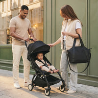Famille parisienne moderne, papa souriant qui pousse une poussette YOYO dépliée avec bébé métis assis dedans et maman aux longs cheveux châtain portant un sac à langer bandoulière en cuir noir sur l'épaule, devanture verte sauge parisienne