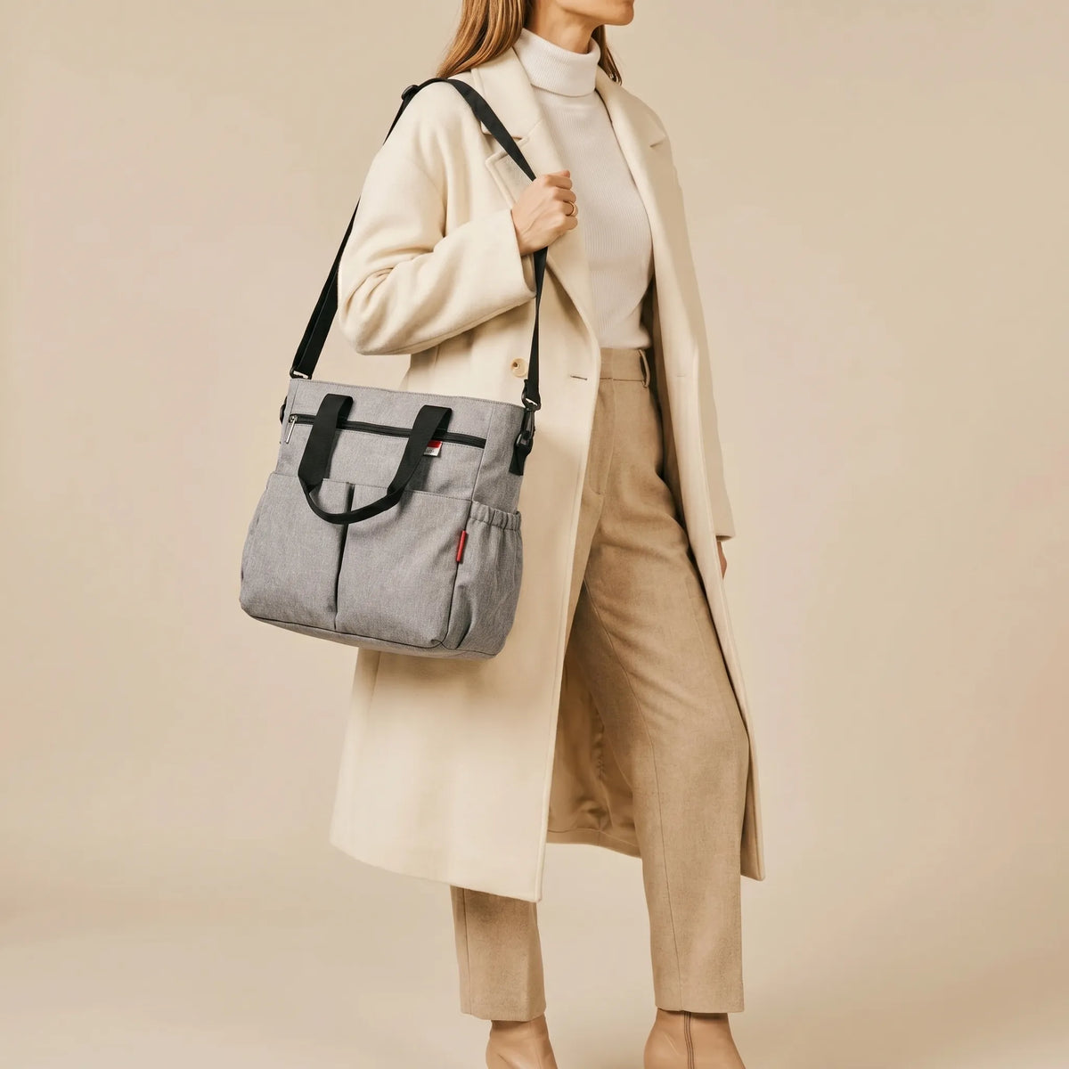 Femme en manteau crème portant un sac à langer bandoulière gris chiné avec poches latérales sur l'épaule