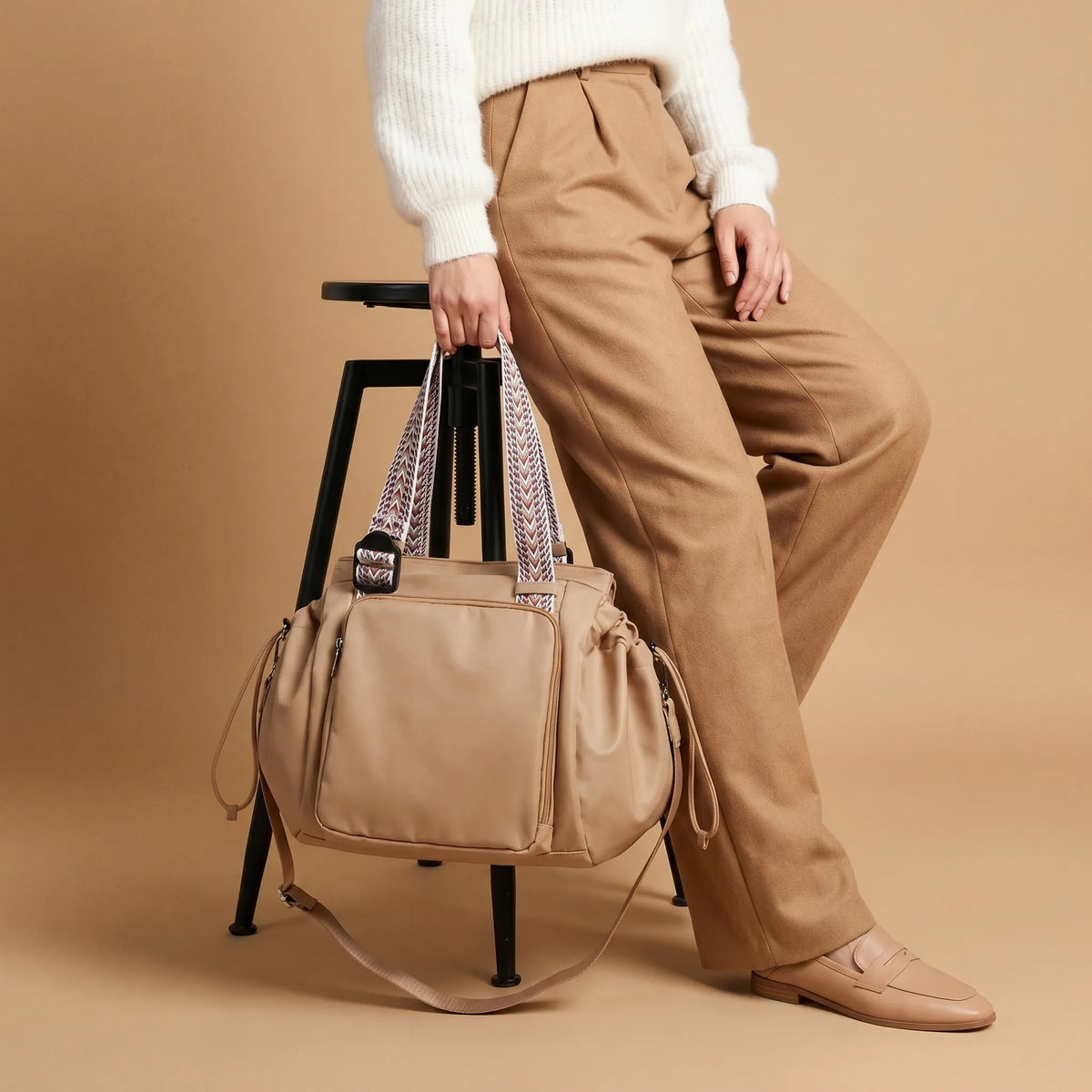 Femme en pantalon caramel et slippers nude tenant un sac à langer beige souple avec anses jacquard tissées à motif chevron multicolore appuyée sur un tabouret