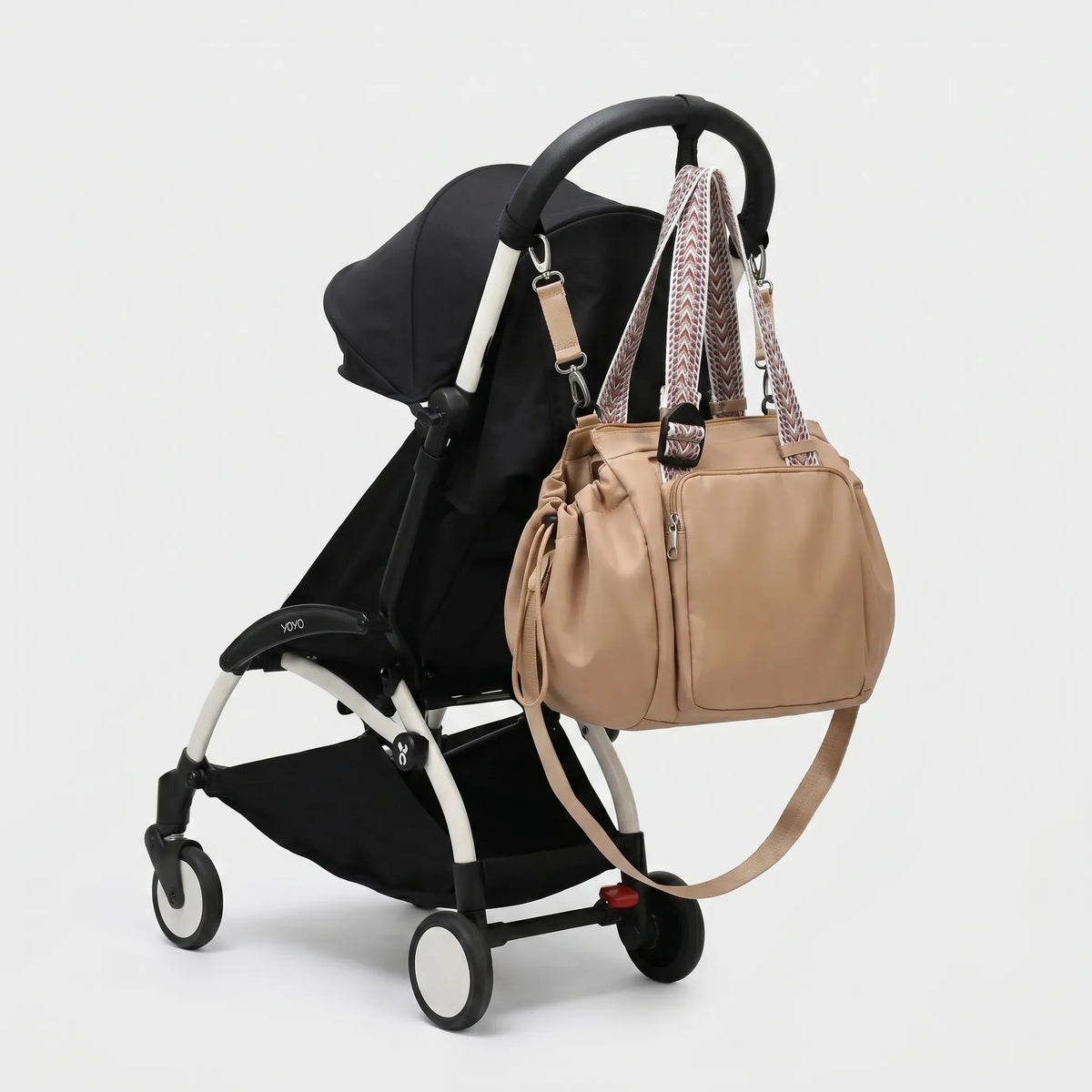 Sac à langer beige nude arrondi avec sangles boho multicolores transformables, 
suspendu au guidon d'une poussette Babyzen YOYO noire — vue lifestyle studio