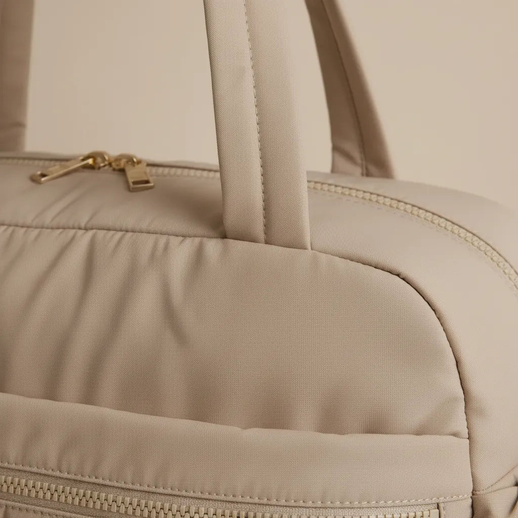 Gros plan sur le haut du sac à langer matelassé beige sable, mettant en valeur la texture tissu structuré rembourrée, la poignée double surpiquée ton sur ton, la fermeture éclair avec curseur doré champagne et les surpiqûres de finition soignées, fond beige rosé épuré ambiance premium

