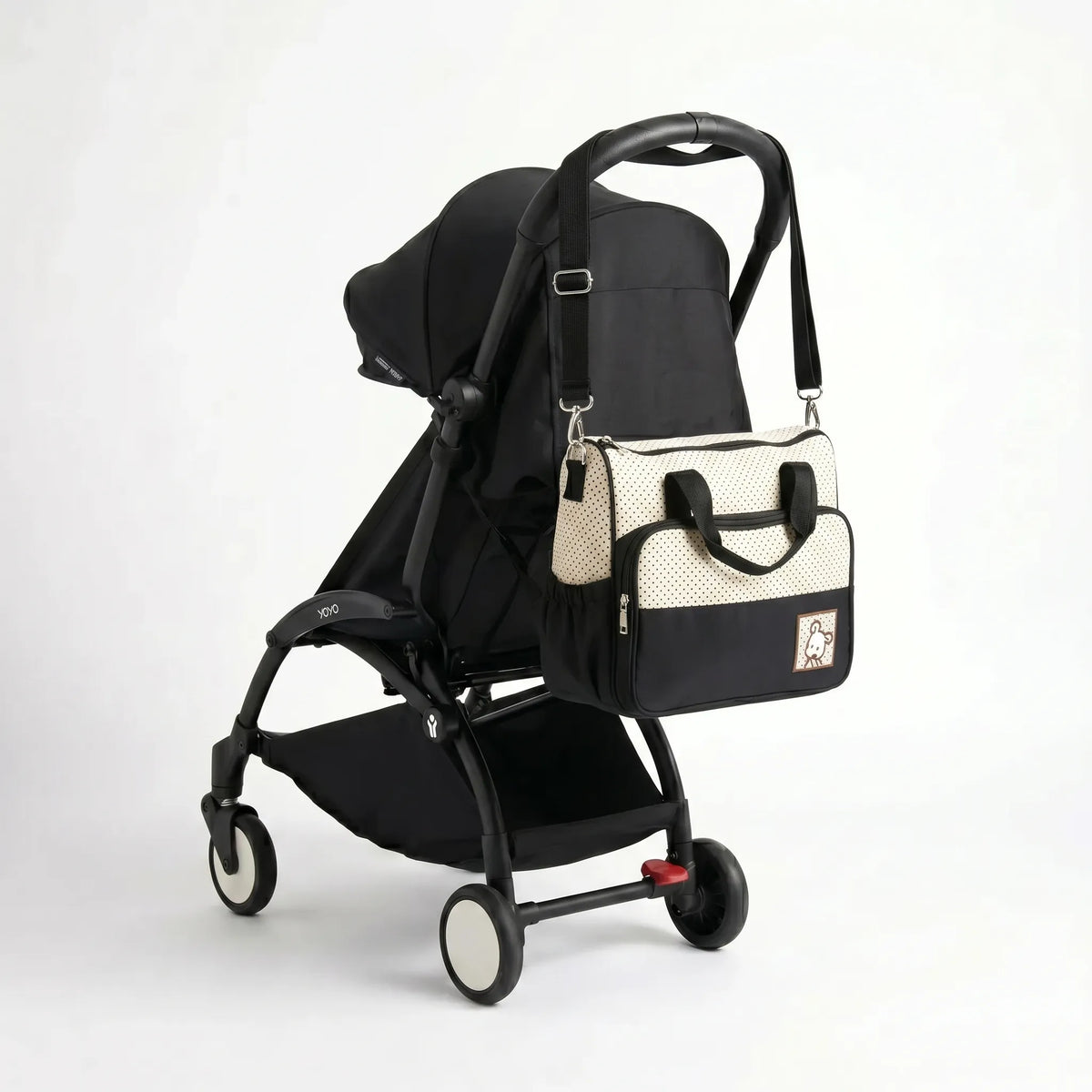 Sac à langer format cabas bicolore écru à pois et noir avec badge ourson et bandoulière accroché au guidon d'une poussette compacte noire de type Yoyo
