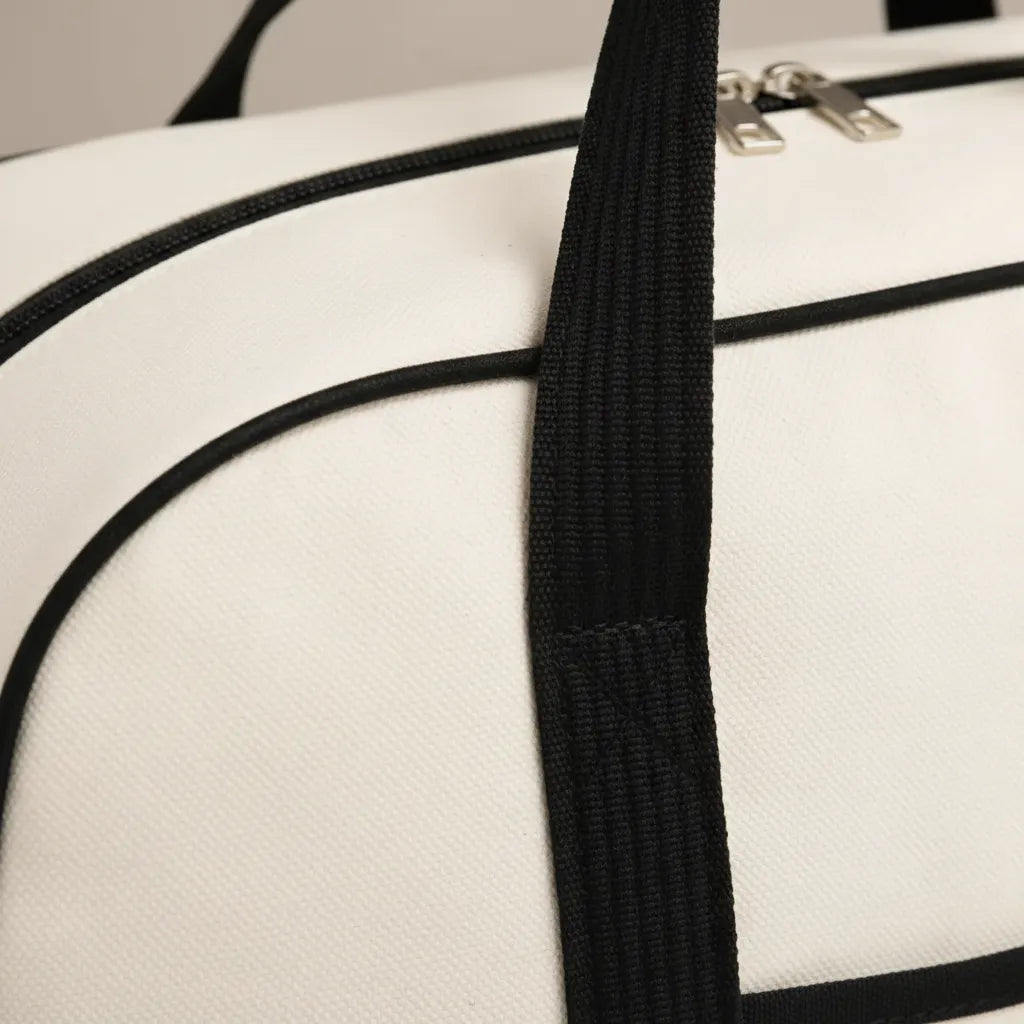 Gros plan sur le flanc supérieur d'un sac à langer bicolore blanc cassé ivoire et noir, révélant la texture canvas tissé serré à grain fin, le passepoil noir mat cousu en bordure de rabat, la large poignée tressée noire avec point de fixation surpiqué renforcé, et la fermeture éclair double curseur argenté brossé visible en haut du cadre, fond studio gris neutre, éclairage rasant valorisant la qualité et la robustesse du textile