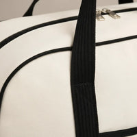 Gros plan sur le flanc supérieur d'un sac à langer bicolore blanc cassé ivoire et noir, révélant la texture canvas tissé serré à grain fin, le passepoil noir mat cousu en bordure de rabat, la large poignée tressée noire avec point de fixation surpiqué renforcé, et la fermeture éclair double curseur argenté brossé visible en haut du cadre, fond studio gris neutre, éclairage rasant valorisant la qualité et la robustesse du textile
