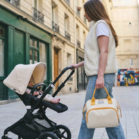 Maman aux longs cheveux châtain marchant dans une rue parisienne en poussant la poussette de son bébé tout en portant à la main un sac à langer crème aux poignées jaunes
