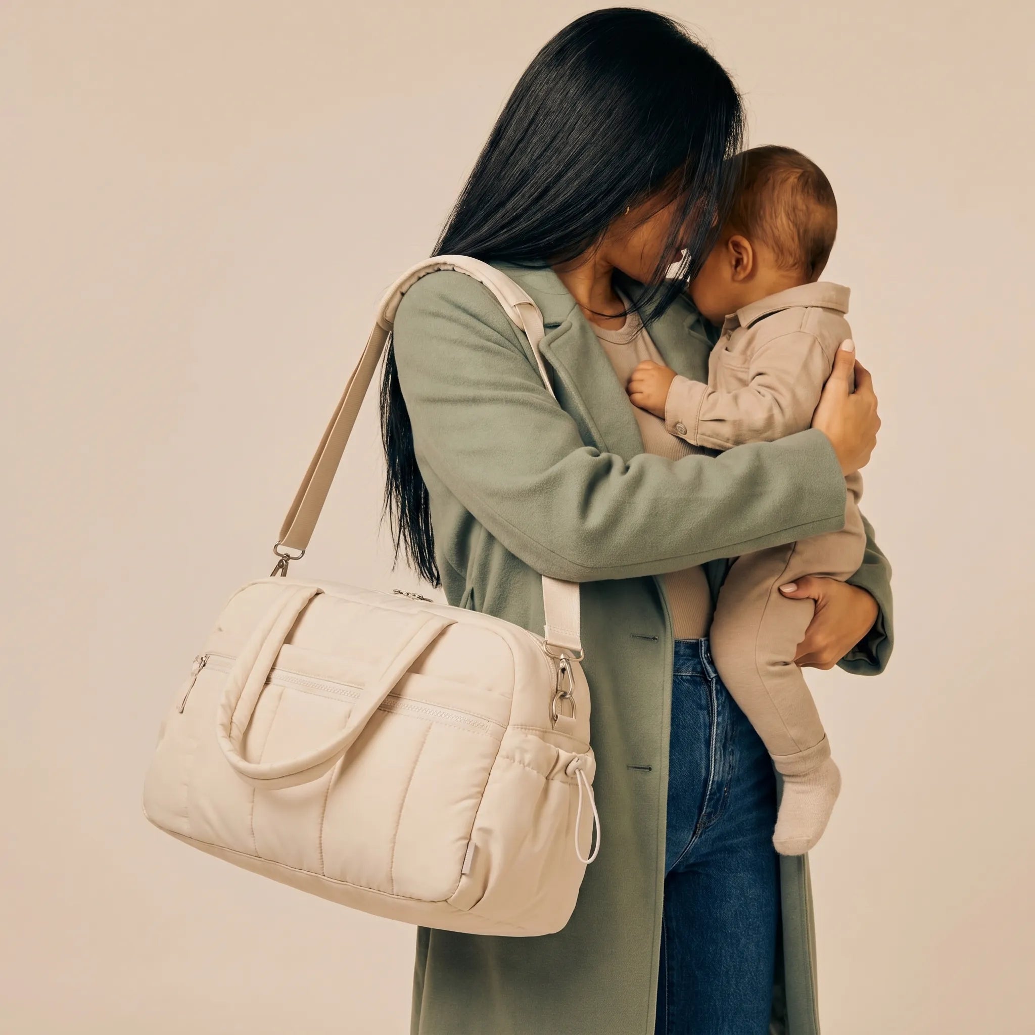 Femme portant un sac à langer duffle beige matelassé en bandoulière tout en tenant son bébé contre son épaule