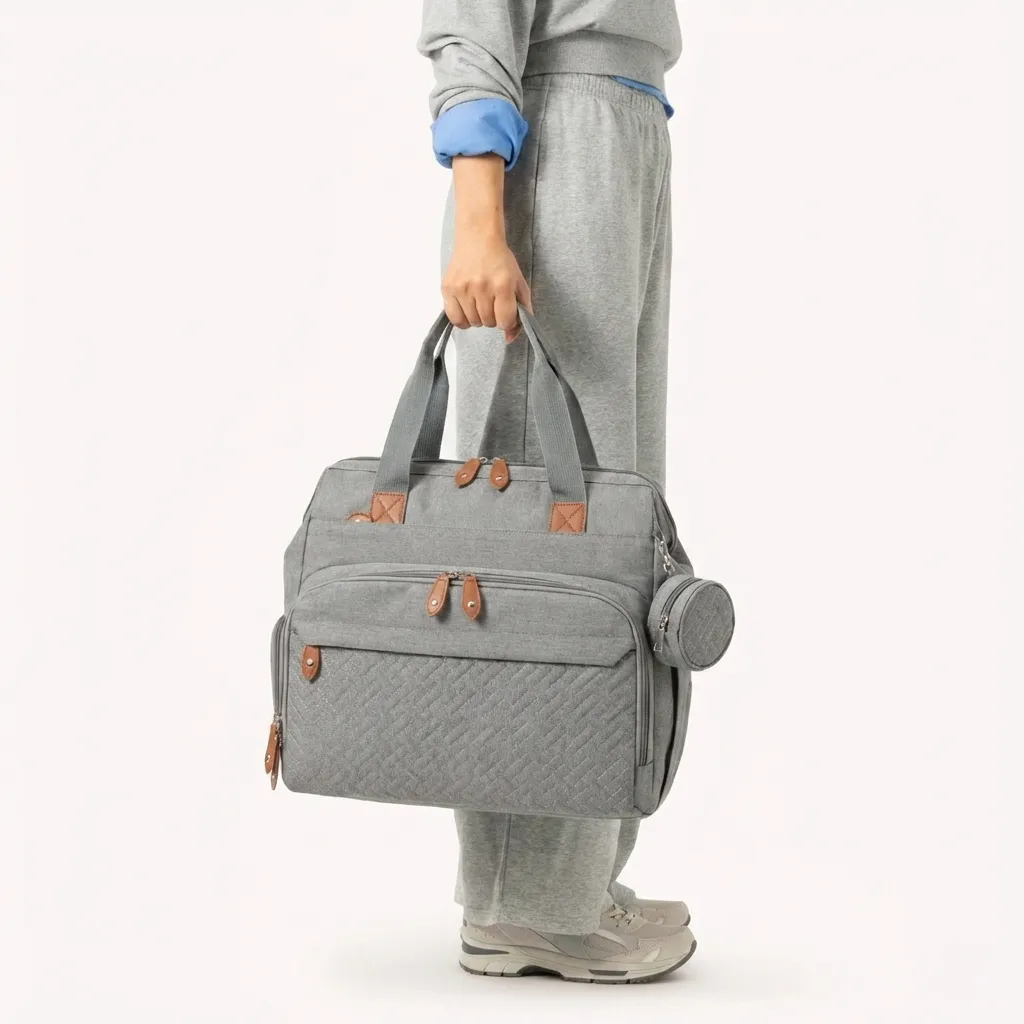 Personne tenant un sac à langer gris grande capacité par les poignées avec détails en cuir marron et pochette ronde sur le côté