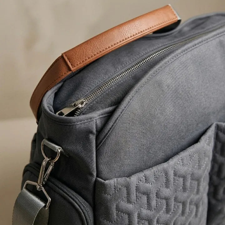 Gros plan sur le sac à langer gris matelassé avec poignée en cuir camel, fermeture éclair argentée et bandoulière réglable – détail coutures et matières haut de gamme