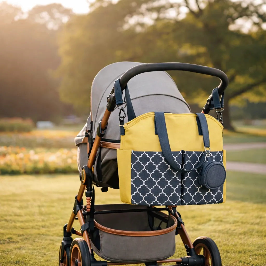 Sac à langer jaune et gris avec motif géométrique fixé à une poussette dans un parc, avec trousse assortie suspendue, ambiance sortie bébé en plein air.