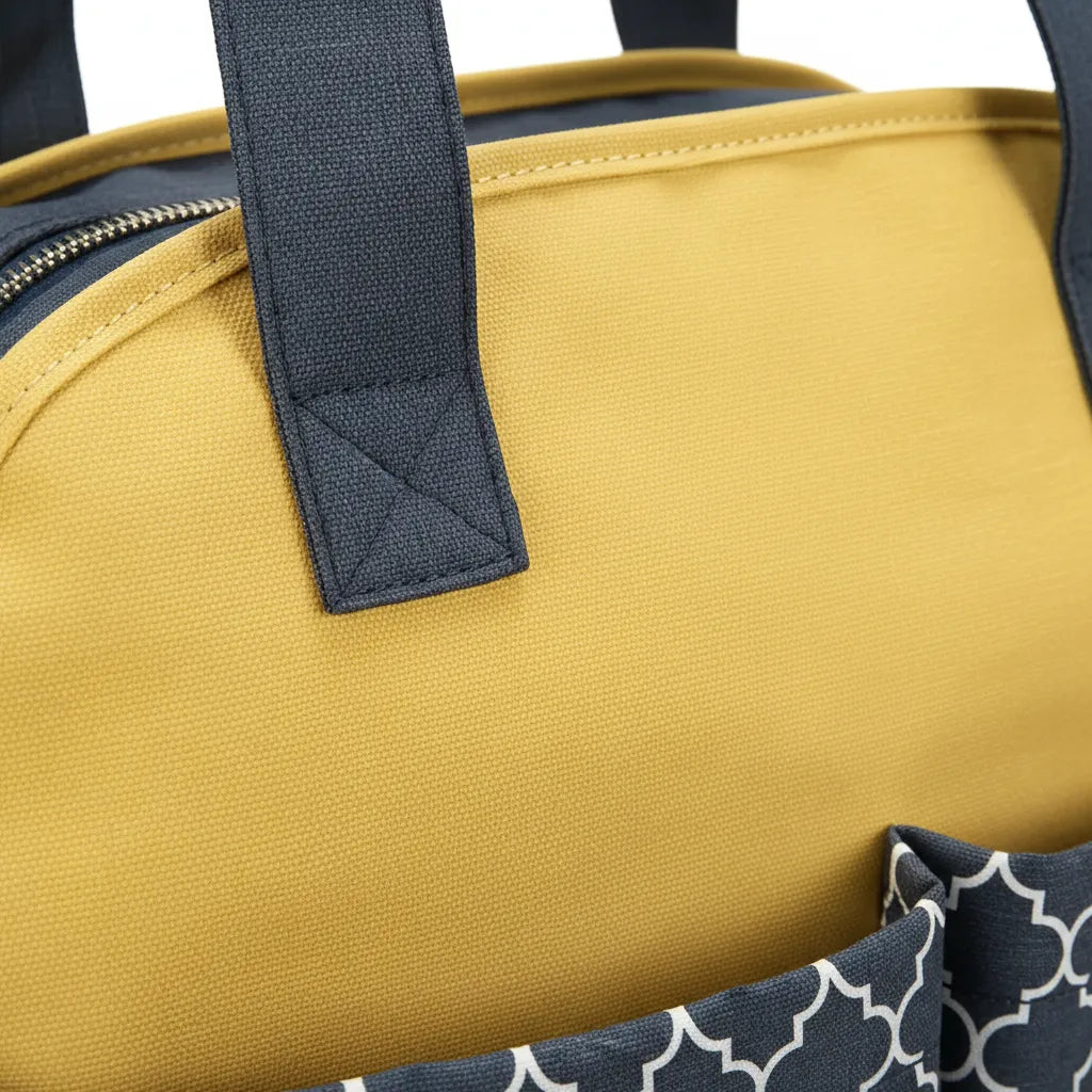 Gros plan sur le flanc supérieur d'un sac à langer bicolore jaune moutarde et gris anthracite, mettant en valeur la texture tissu canvas tissé serré, la poignée large gris ardoise avec renfort triangulaire surpiqué double point au niveau de l'attache, la fermeture éclair argent brossé sur le bord supérieur et le motif treillis géométrique blanc visible en bas du cadre, fond studio neutre éclairage rasant révélant la qualité du textile