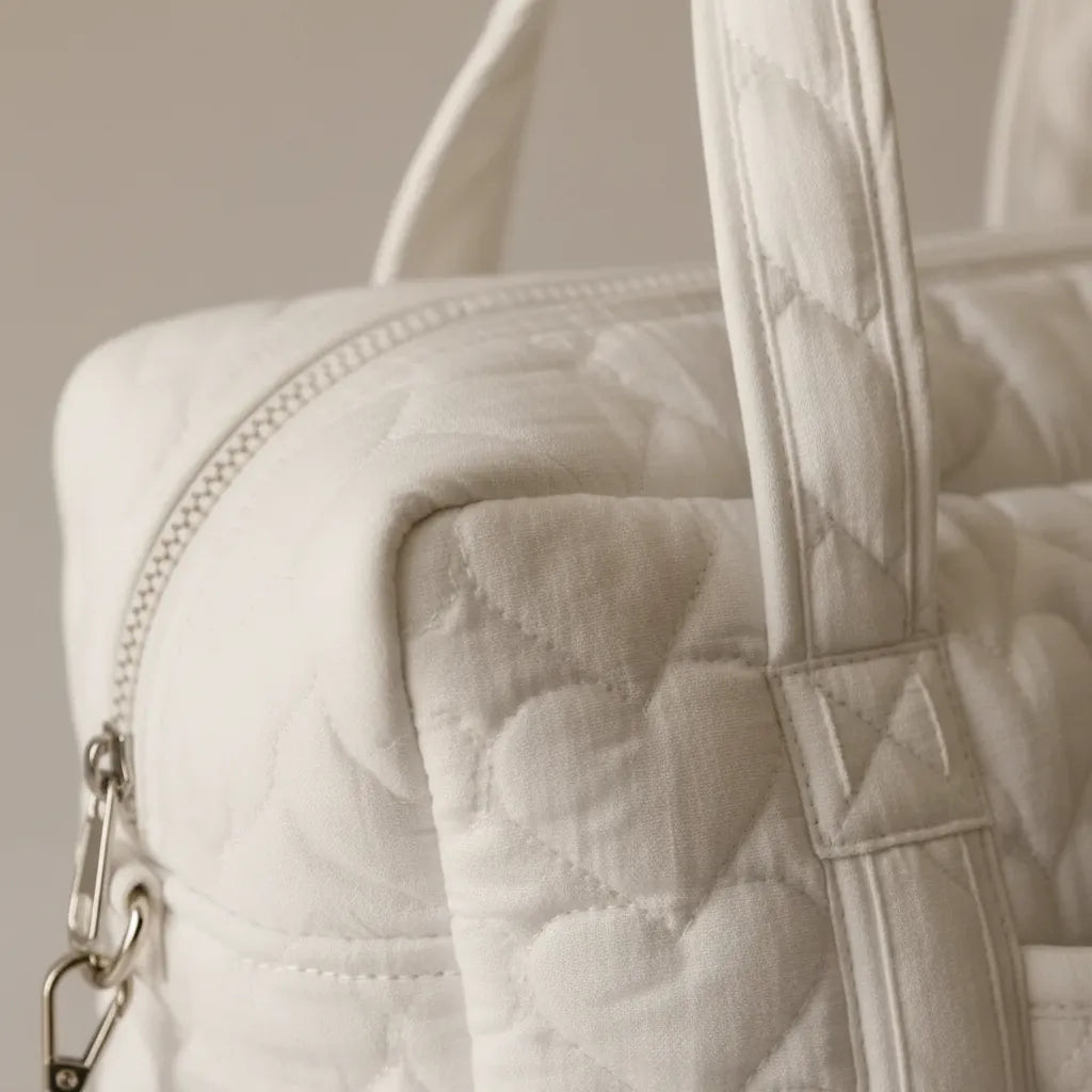 Gros plan sur la texture gaze de coton matelassée blanc ivoire d'un sac à langer, révélant les piqûres de matelassage en relief, la fermeture éclair blanche, la poignée rembourrée et le mousqueton argenté de la bandoulière détachable