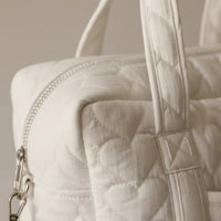 Gros plan sur la texture gaze de coton matelassée blanc ivoire d'un sac à langer, révélant les piqûres de matelassage en relief, la fermeture éclair blanche, la poignée rembourrée et le mousqueton argenté de la bandoulière détachable