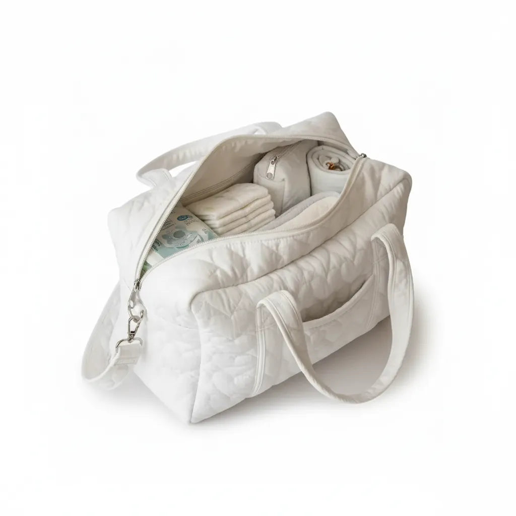 Vue intérieure d'un sac à langer matelassé blanc ivoire grand ouvert sur fond blanc, révélant un compartiment principal bien organisé avec couches pliées, lange roulé beige, pochette intérieure zippée et lingettes bébé, double poignée rembourrée et mousqueton bandoulière argenté visible sur le côté