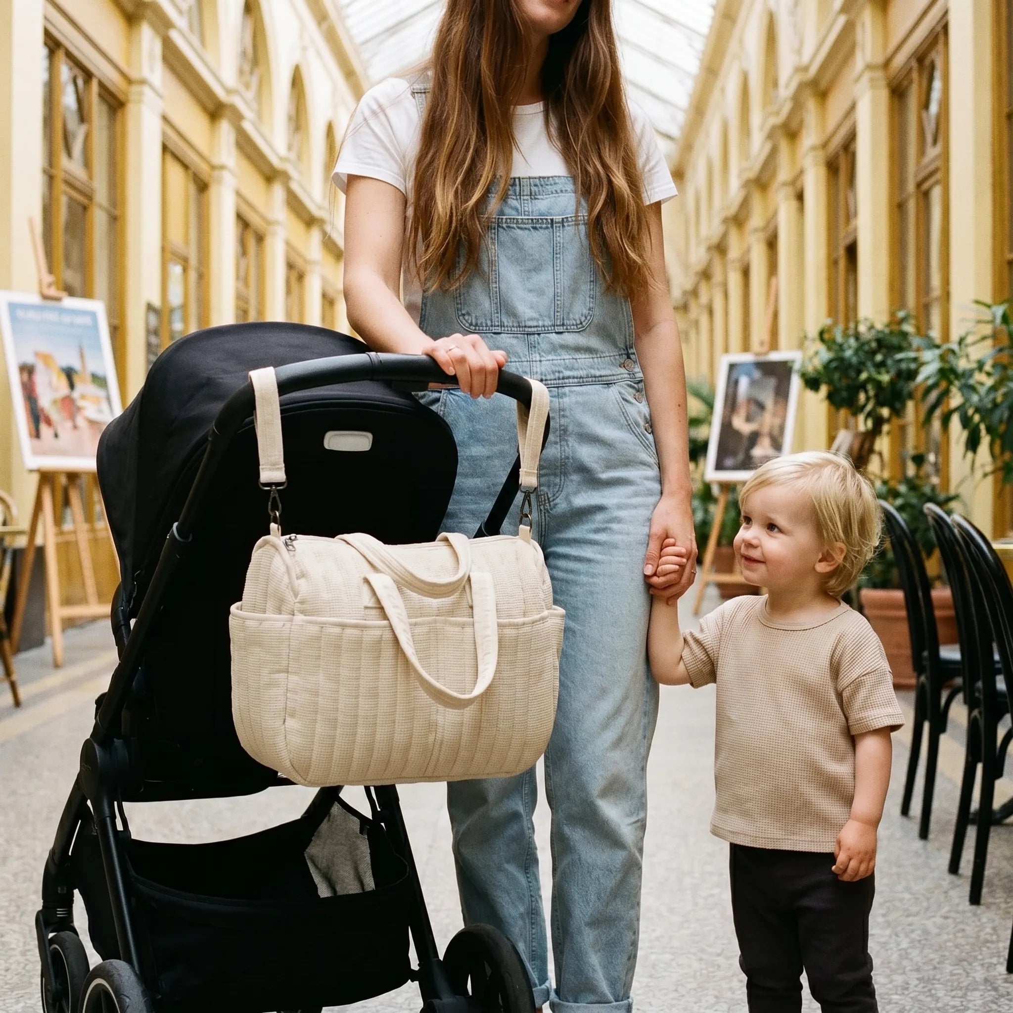Maman aux longs cheveux châtain en salopette tenant la main de son jeune enfant blond avec un sac à langer matelassé crème accroché à la poussette dans un passage couvert parisien