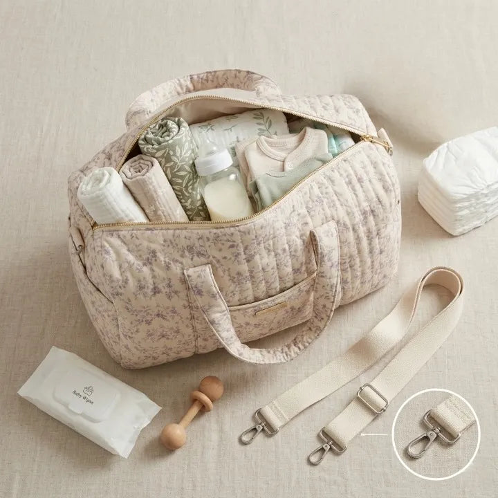 Vue du dessus d'un sac à langer matelassé beige à fleurs mauves ouvert, révélant son grand compartiment organisé : lange mousseline, biberon, body bébé vert sauge, couverture florale, lingettes et couches posées à côté, hochet en bois, avec zoom sur la bandoulière beige à mousquetons argentés détachables