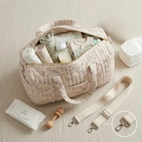 Vue du dessus d'un sac à langer matelassé beige à fleurs mauves ouvert, révélant son grand compartiment organisé : lange mousseline, biberon, body bébé vert sauge, couverture florale, lingettes et couches posées à côté, hochet en bois, avec zoom sur la bandoulière beige à mousquetons argentés détachables