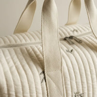 Gros plan sur la texture matelassée côtelée ivoire d'un sac à langer, mettant en valeur les broderies brindilles grises, la poignée en coton tressé beige renforcée, la fermeture éclair double curseur argentée et la qualité des surpiqûres parallèles