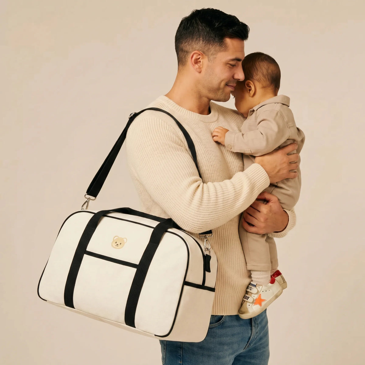 Papa portant son bébé contre lui avec un sac à langer de maternité en toile écrue, sangles noires et logo ourson doré, porté à l'épaule