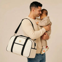 Papa portant son bébé contre lui avec un sac à langer de maternité en toile écrue, sangles noires et logo ourson doré, porté à l'épaule
