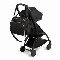 Sac à langer noir matelassé avec zips dorés, bandoulière et poches latérales accroché au guidon d'une poussette compacte noire de type Yoyo