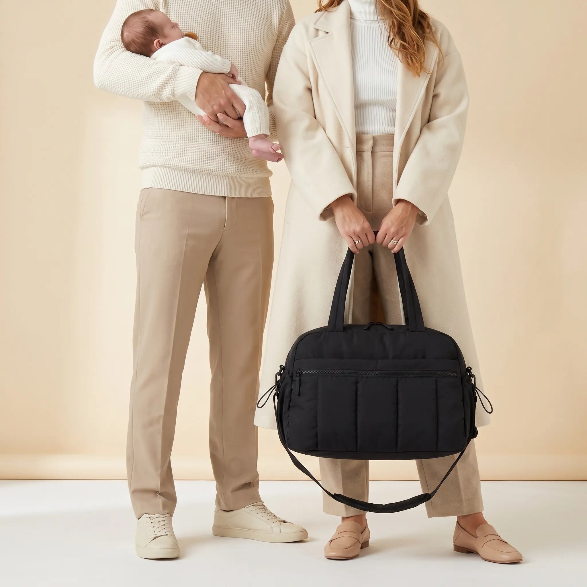 Couple de parents en tenue beige et crème quiet luxury tenant un sac à langer noir matelassé avec leur nouveau-né endormi sur fond beige studio