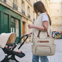 Maman aux longs cheveux châtain en sherpa crème poussant la poussette de son bébé dans une rue parisienne avec un sac à langer rayé écru aux anses cuir cognac sur l'épaule