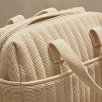 Gros plan sur la texture matelassée côtelée beige d'un sac à langer maternité solide imperméable : anses doubles renforcées cousues solidement, fermeture éclair robuste et poches latérales extérieures accès rapide