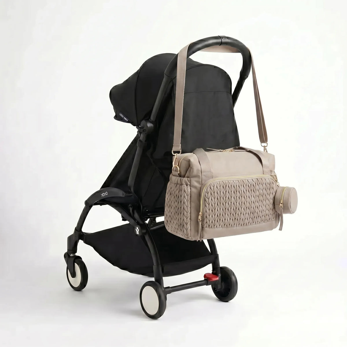 Sac à langer format week-end taupe matelassé effet torsadé avec zips dorés et pochette ronde assortie accroché au guidon d'une poussette compacte noire de type Yoyo