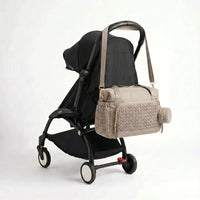 Sac à langer format week-end taupe matelassé effet torsadé avec zips dorés et pochette ronde assortie accroché au guidon d'une poussette compacte noire de type Yoyo
