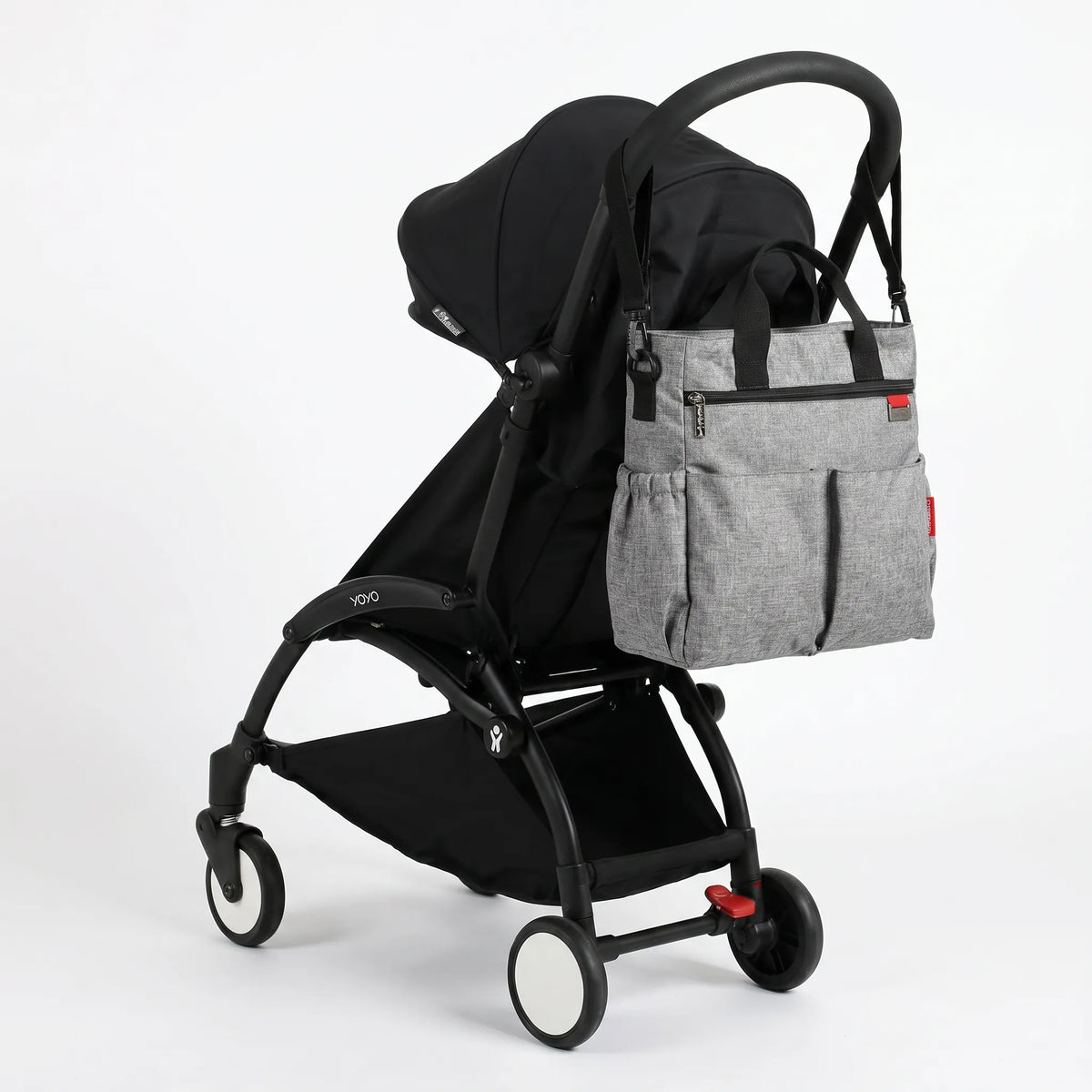 Sac à langer cabas gris chiné avec poignées noires et double poche frontale, 
suspendu au guidon d'une poussette Babyzen YOYO noire — vue lifestyle studio