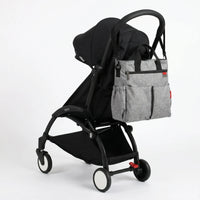 Sac à langer cabas gris chiné avec poignées noires et double poche frontale, 
suspendu au guidon d'une poussette Babyzen YOYO noire — vue lifestyle studio